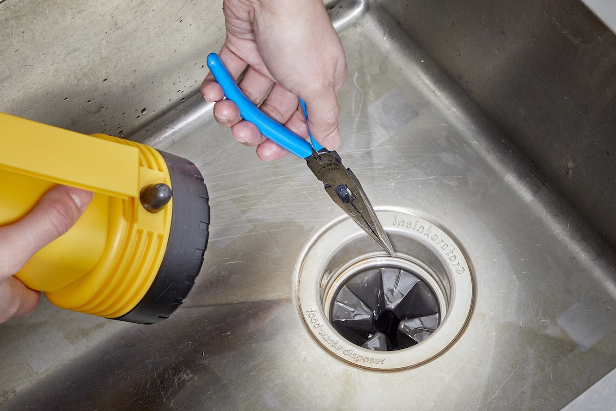 Person fishes needle-nose pliers into garbage disposal while holding a flashlight nearby.