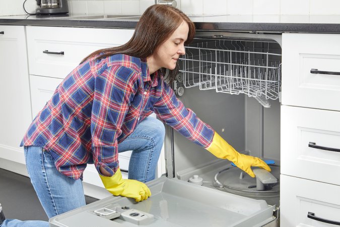 Woman wearing a flannel shirt and cleaning gloves reaching into her dishwasher to access the filter.
