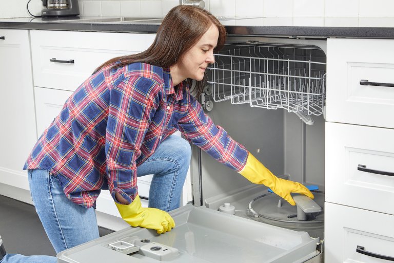 Woman wearing a flannel shirt and cleaning gloves reaching into her dishwasher to access the filter.