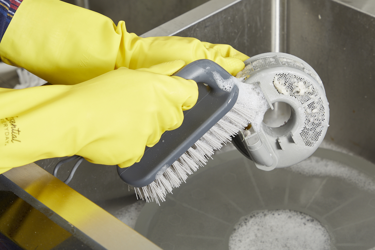 Woman scrubs sudsy dishwasher filter using a scrub brush.