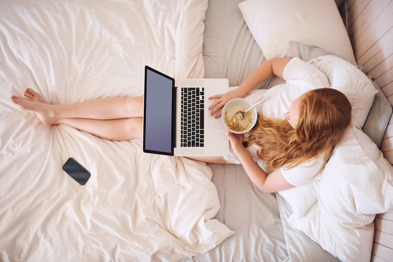 overhead shot of woman in bed with white sheets and bedding, sitting with laptop on lap and eating a bowl of cereal