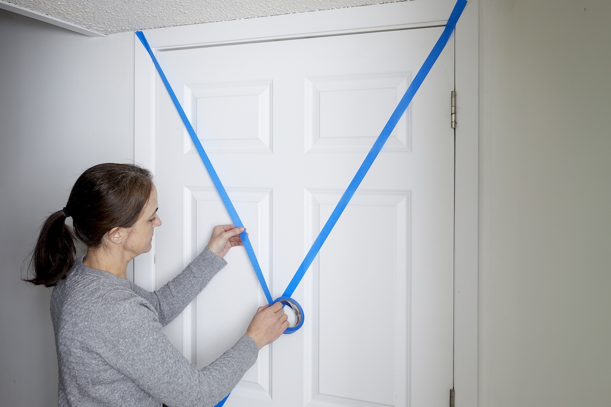 Woman uses painter's tape to make an "X" in tape over a white interior door.