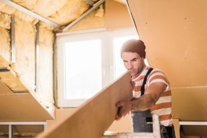 Young man checking plank on construction site under dormer window with exposed insulation.