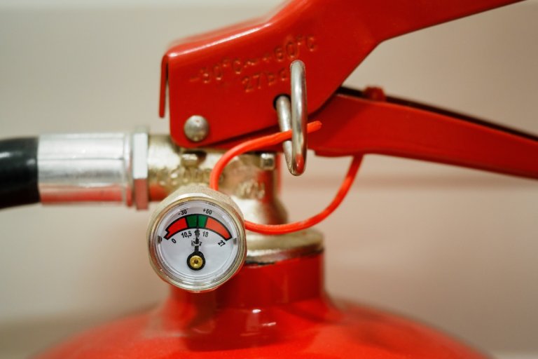 Close up view of the top of a red fire extinguisher showing the nozzle and pressure gauge.