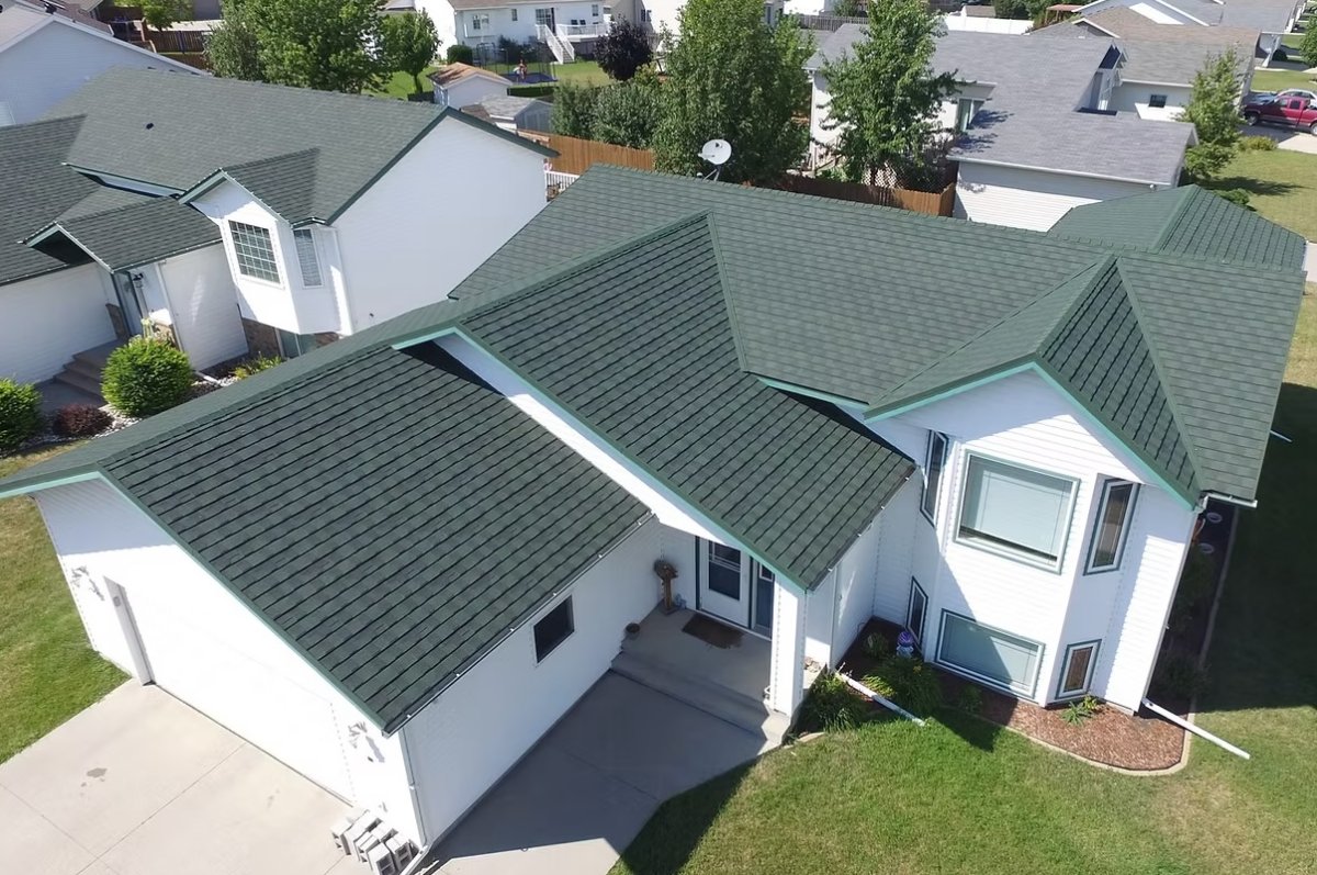Overhead view of a white house with a green metal shingled roof, and a beige concrete driveway in a sunny climate.