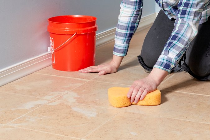 Woman kneels on tile floor sponging floor next to orange bucket.