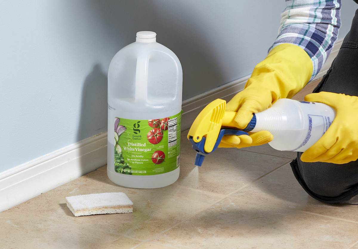 Woman wearing rubber gloves sprays tile floor, a jug of vinegar in background.