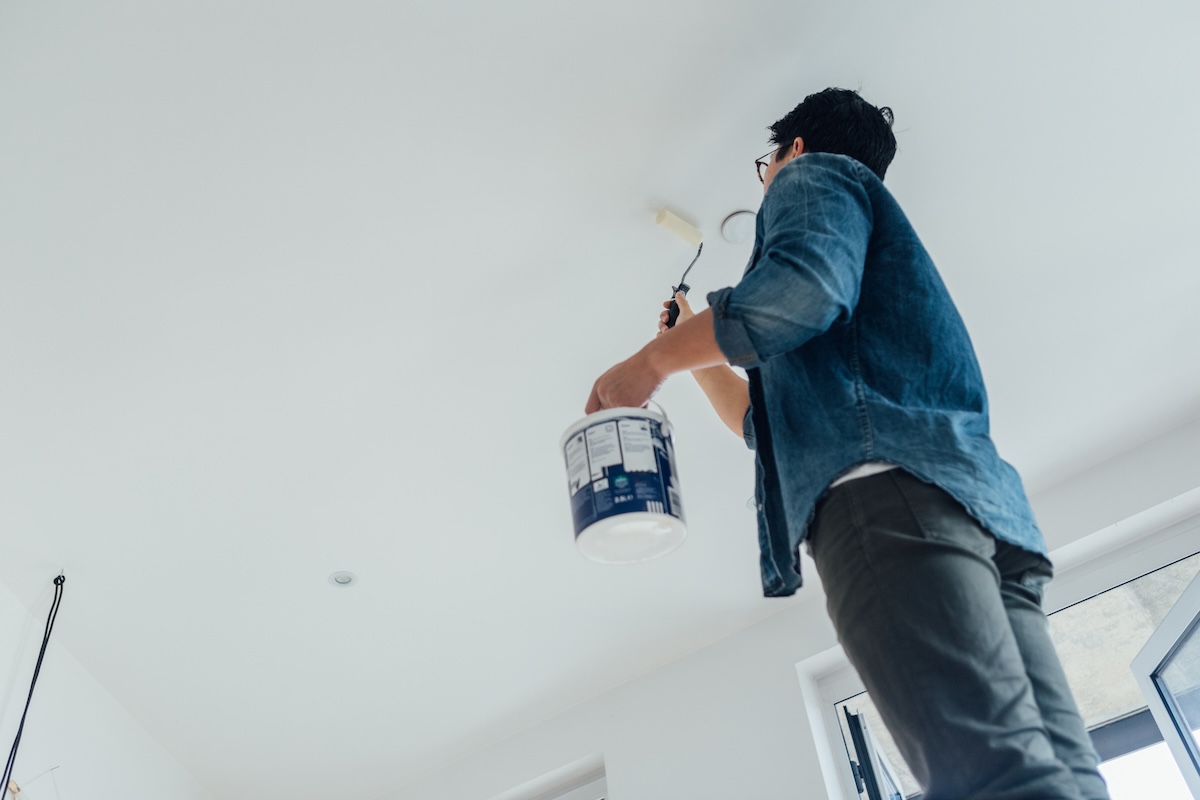 A man in a denim shirt paints the ceiling white using a paint roller.
