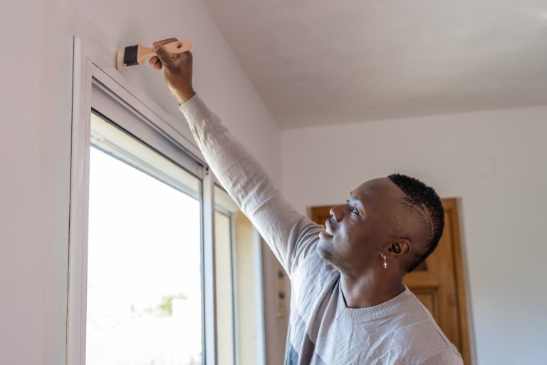 A smiling younger man paints wall trim with white paint.