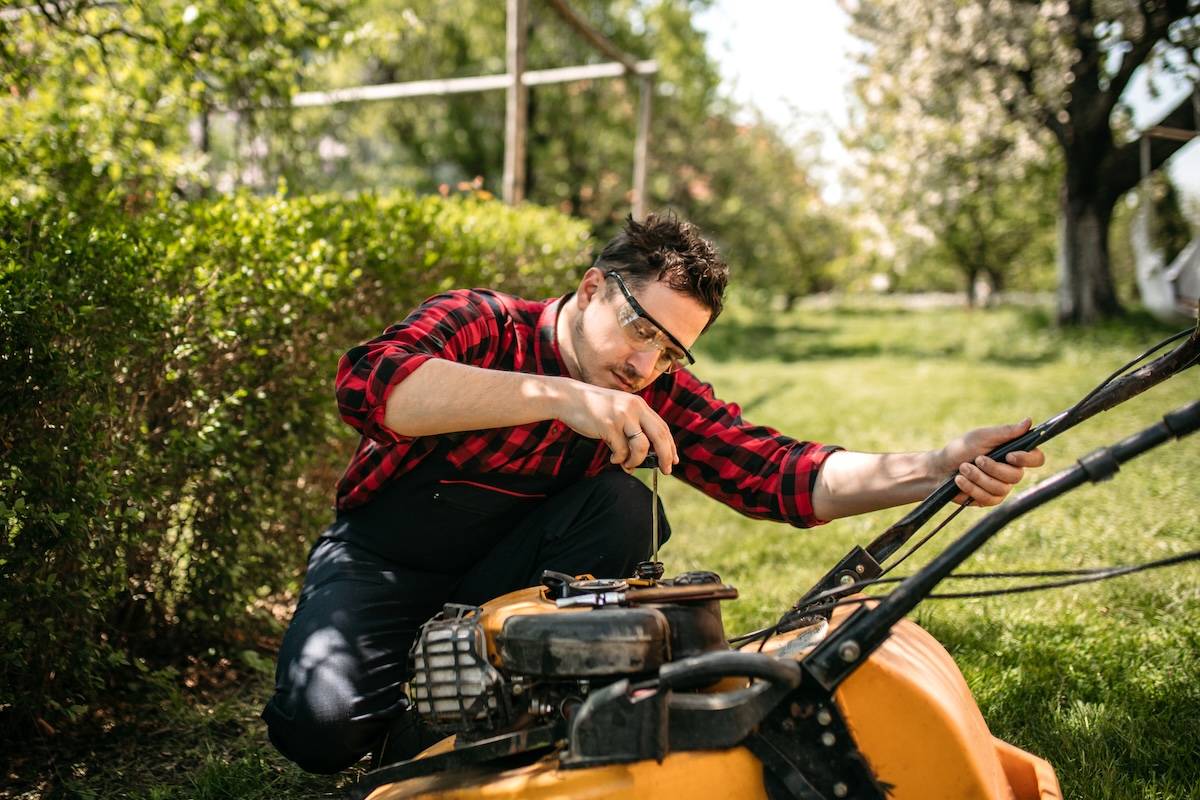 A man in a plaid shirt and safety goggles examines the oil valve of a yellow lawn mower.