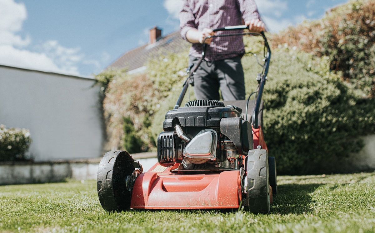 A close up of the engine on a red lawn mower while a man pushes it across the lawn.