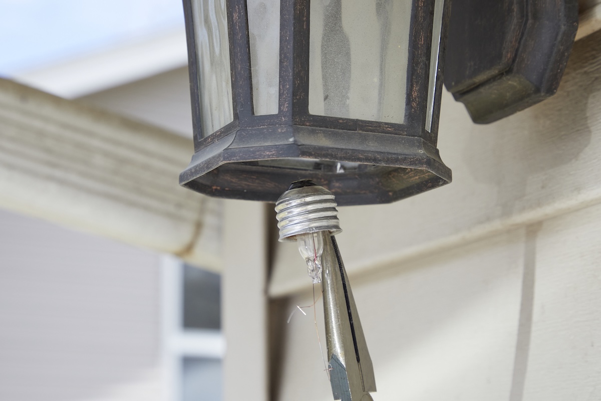 Person uses needle-nose pliers to pull a broken light bulb out from an outdoor light sconce.