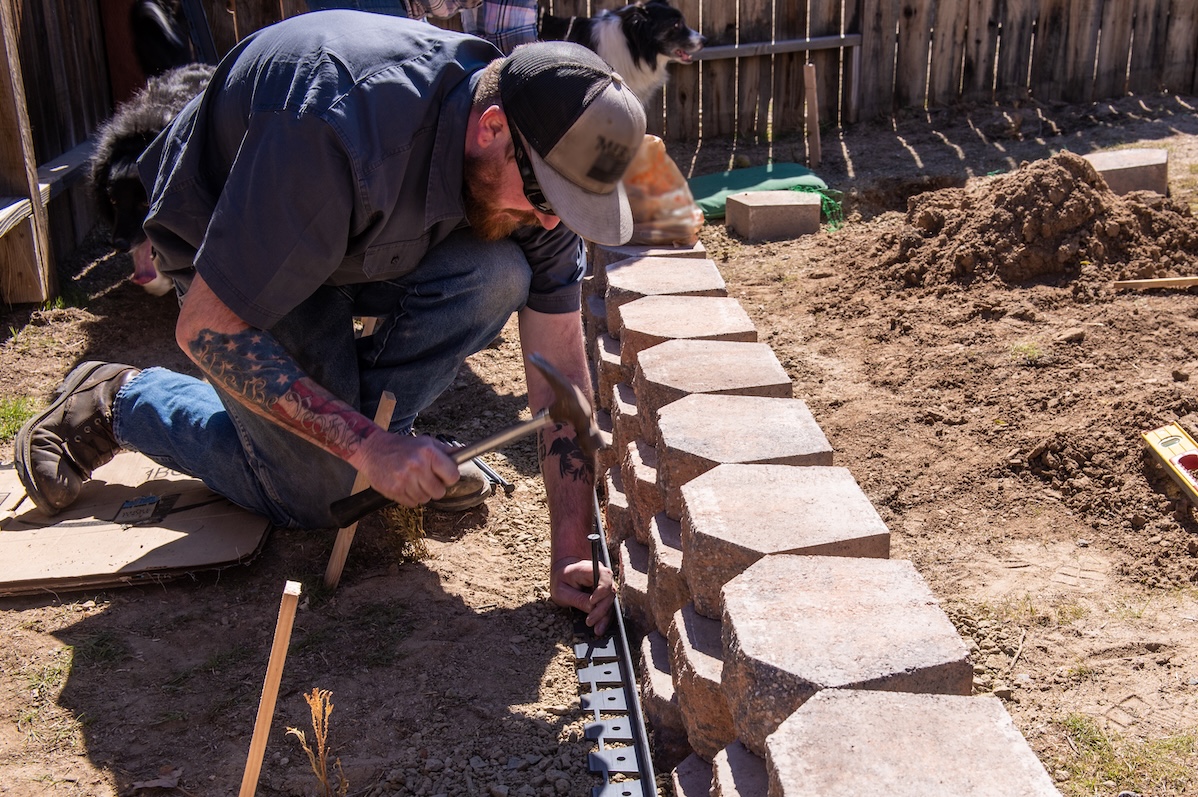 Man with a trucker hat and tattooed arms is building a retaining wall out of paver stones in a yard with no grass.