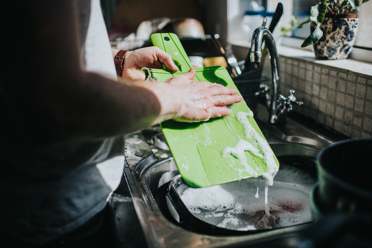 A man washes a green plastic cutting board in the sink.