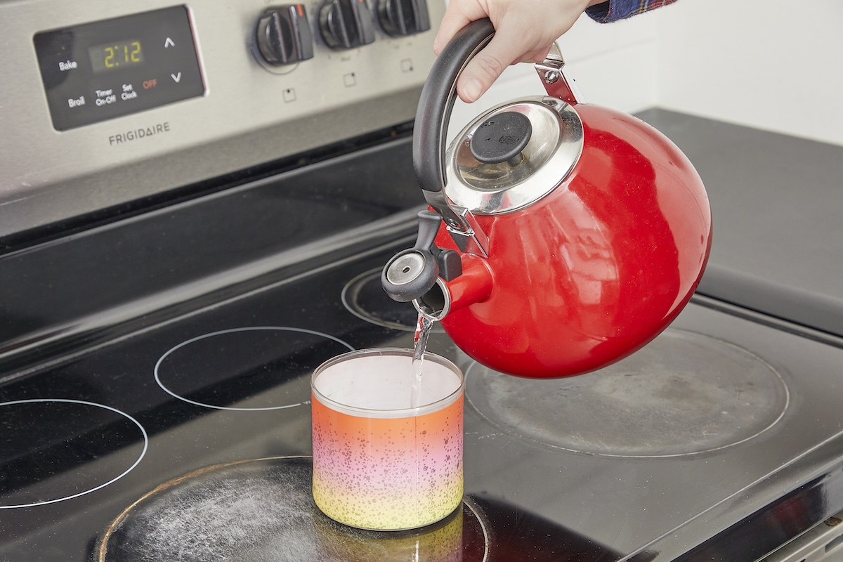 A person pours hot water from a red kettle into a candle jar.