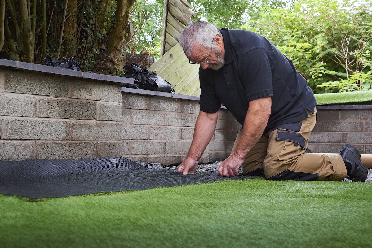 A garden worker lays a patch of landscaping fabric to fit the border of a grass lawn.
