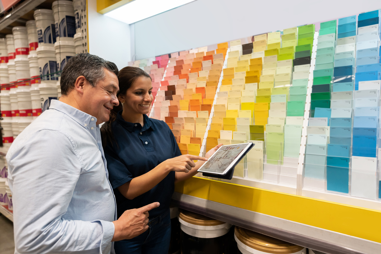 Salesperson in a paint store shows a man something on a tablet, in front of a display of paint chips.