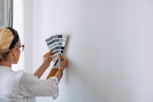 Woman with a headband in her hair holding paint swatches to a white wall.