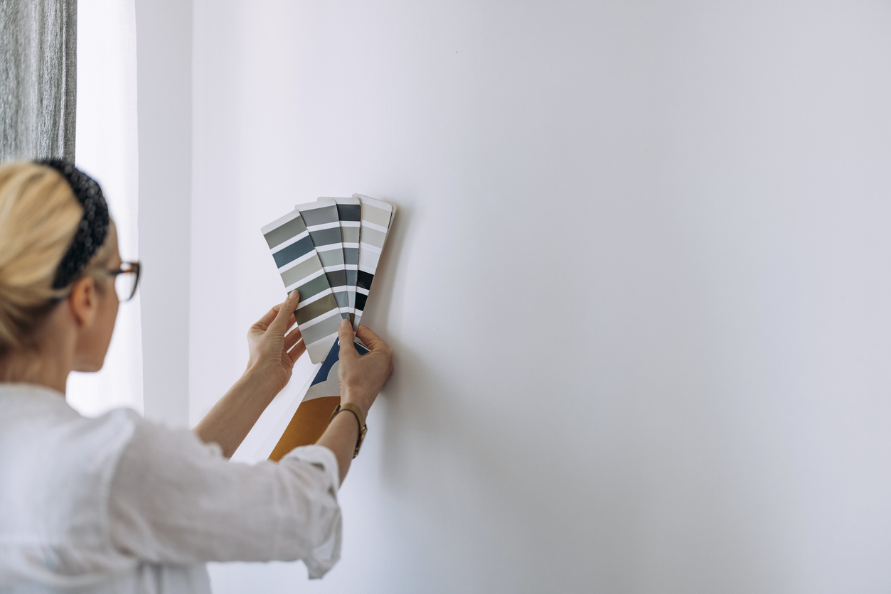 Woman with a headband in her hair holding paint swatches to a white wall.