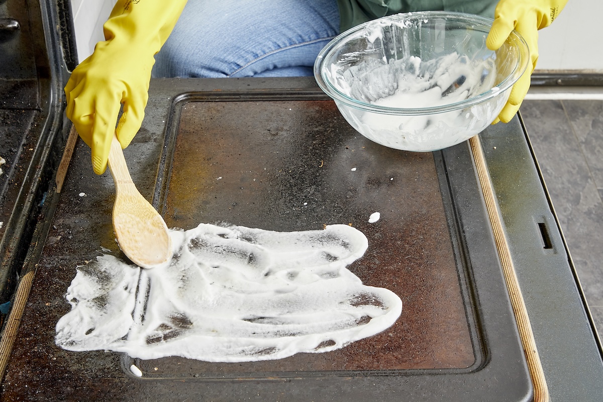Person wearing yellow rubber gloves spreads white cleaning paste from a glass bowl onto oven glass.