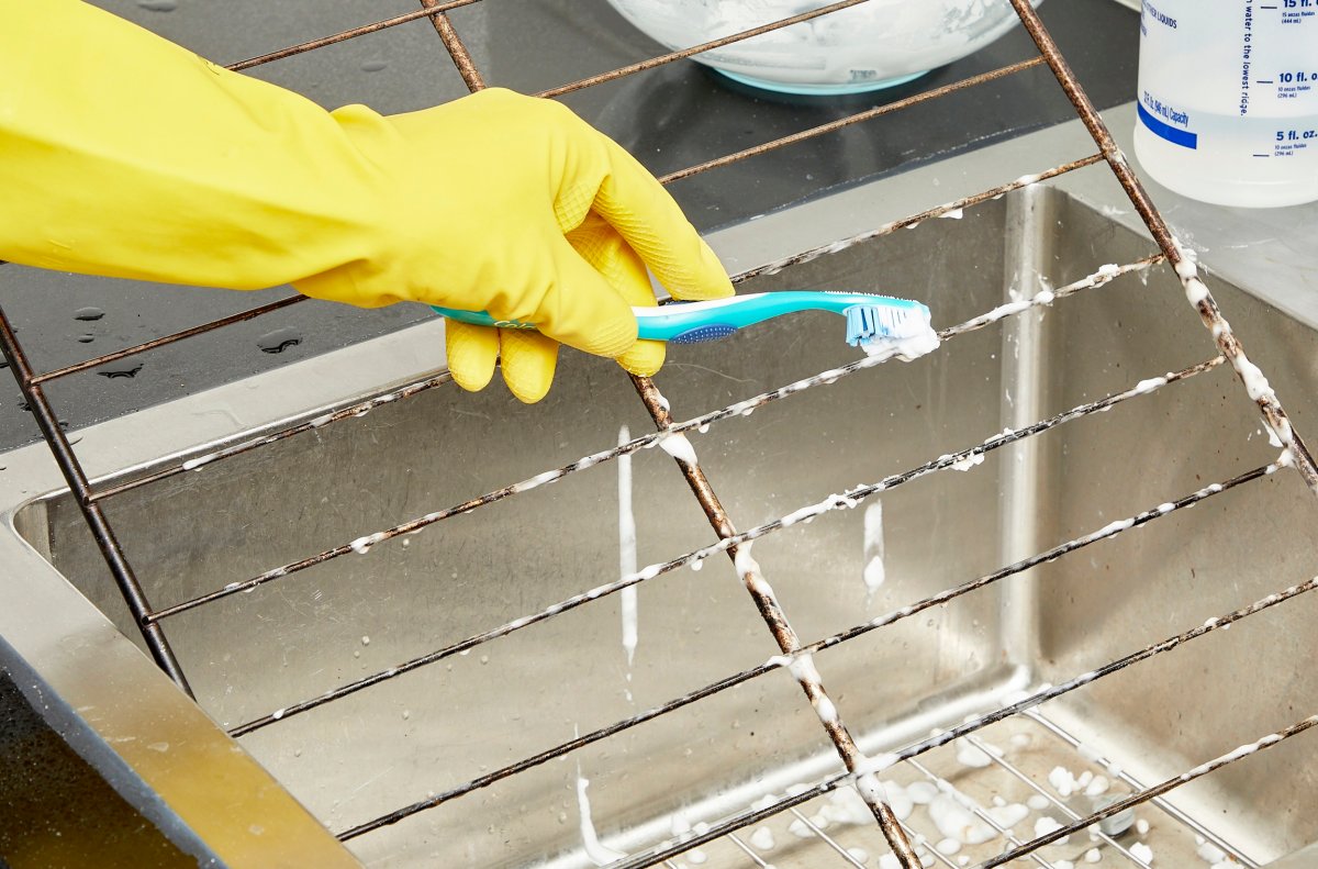Person wearing yellow rubber gloves scrubs dirty oven racks in a stainless sink with a toothbrush.