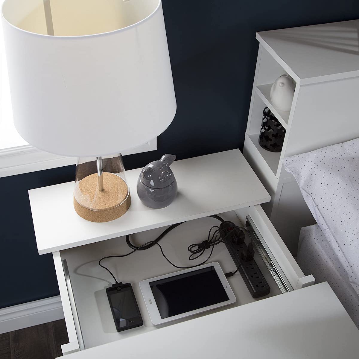 White nightstand with lamp on top; open nightstand drawer reveals phone and tablet plugged into a power strip.