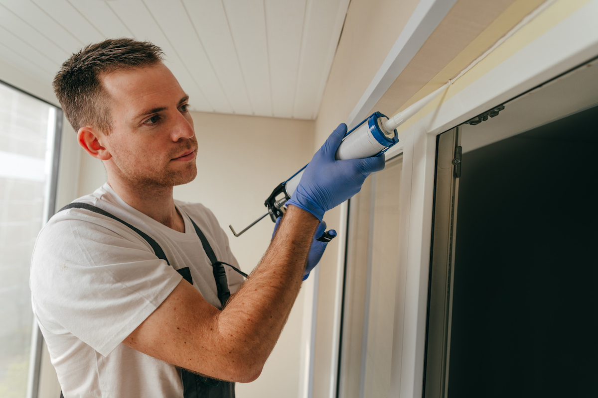 A man is using painter's tape along the seam of an exterior window to help keep his caulking gun straight.
