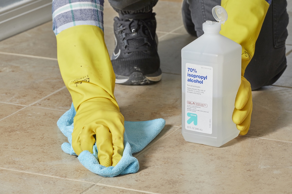 Woman wearing yellow rubber gloves wipes floor with blue towel and rubbing alcohol.