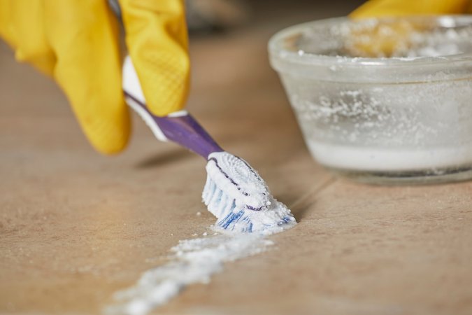 Person wearing yellow rubber gloves cleans tile grout with a toothbrush and a white powdery scrub.