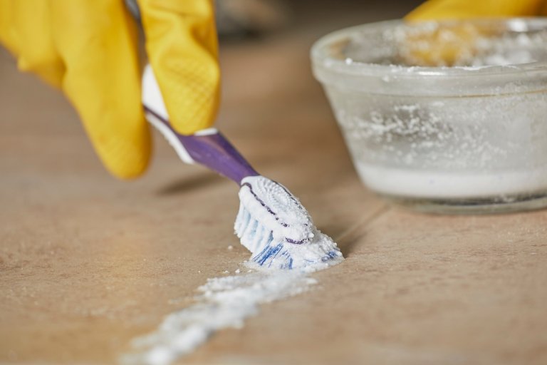 Person wearing yellow rubber gloves cleans tile grout with a toothbrush and a white powdery scrub.