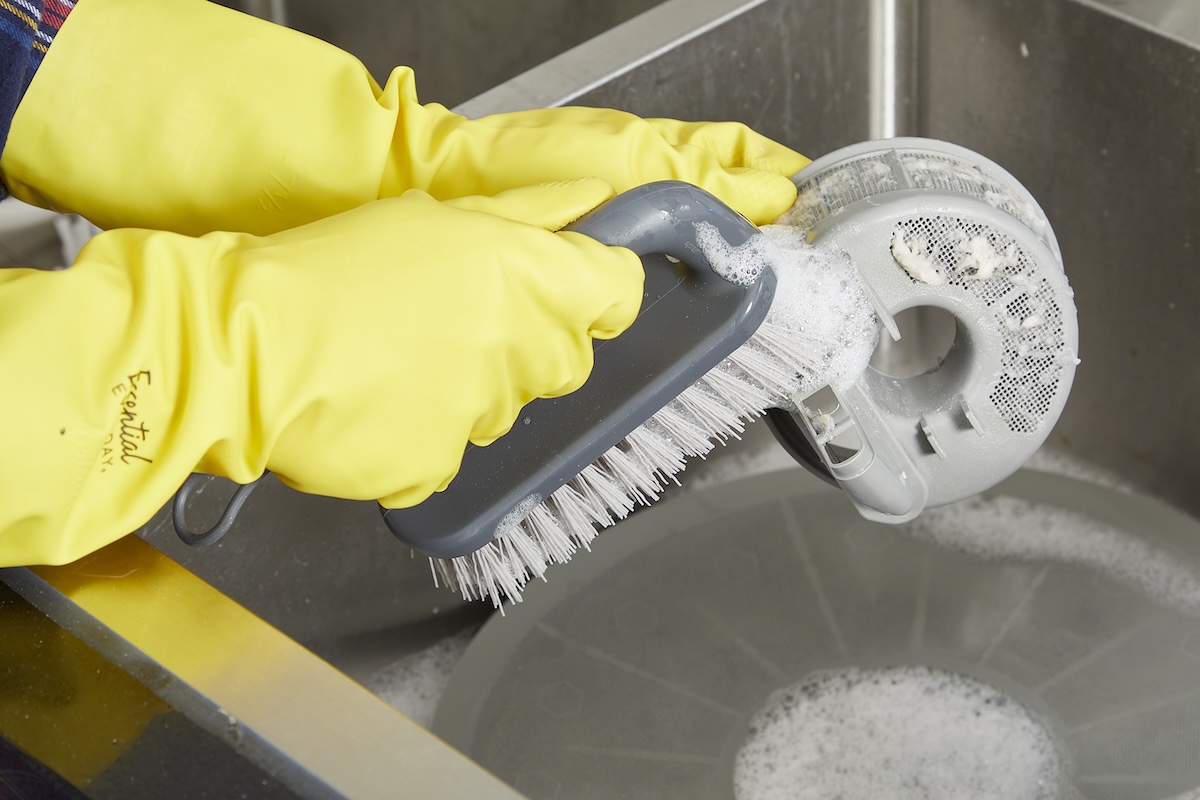Hands with yellow rubber gloves scrub a dishwasher filter with a scrub brush.