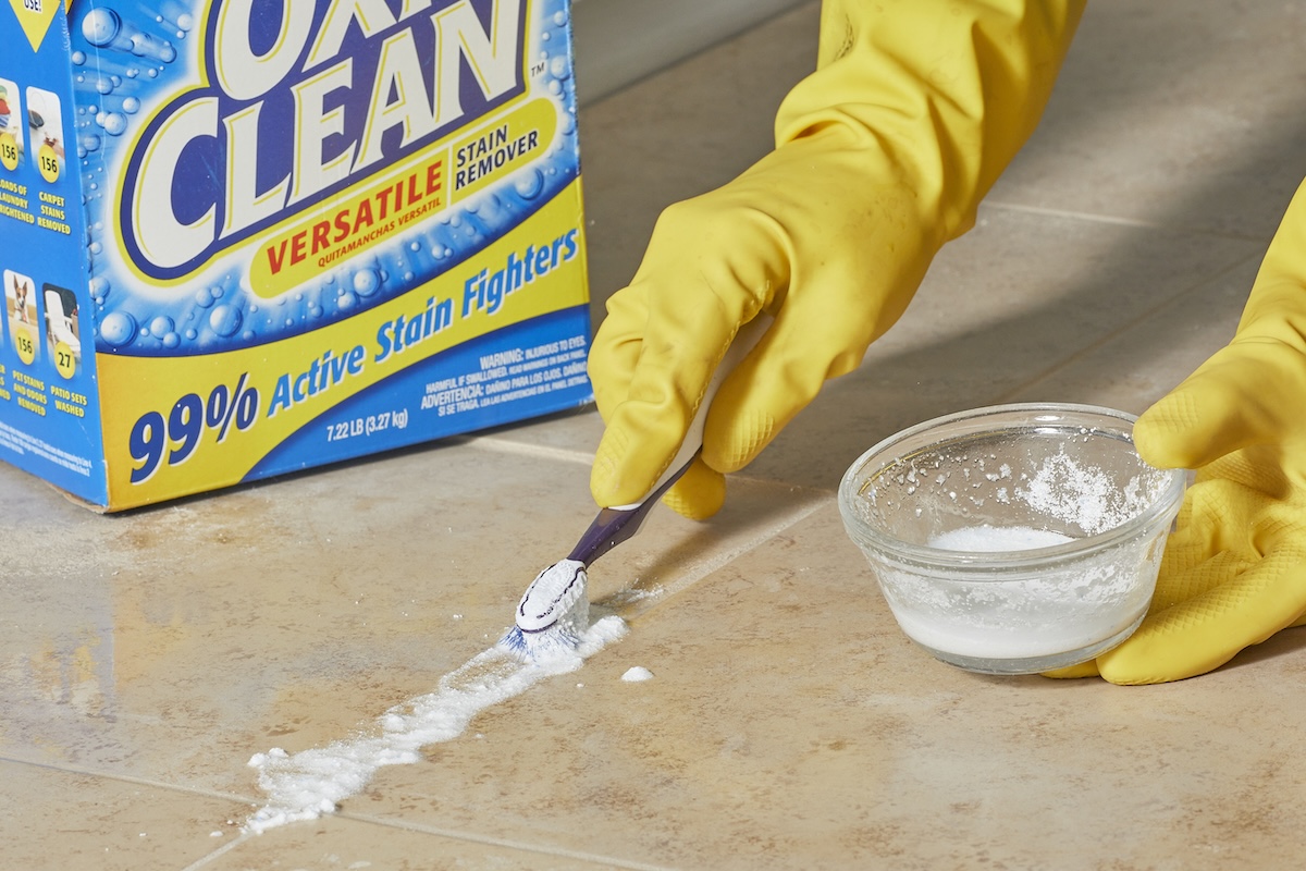 A person wearing yellow, rubber gloves uses a toothbrush to scrub the grout between tiles. 