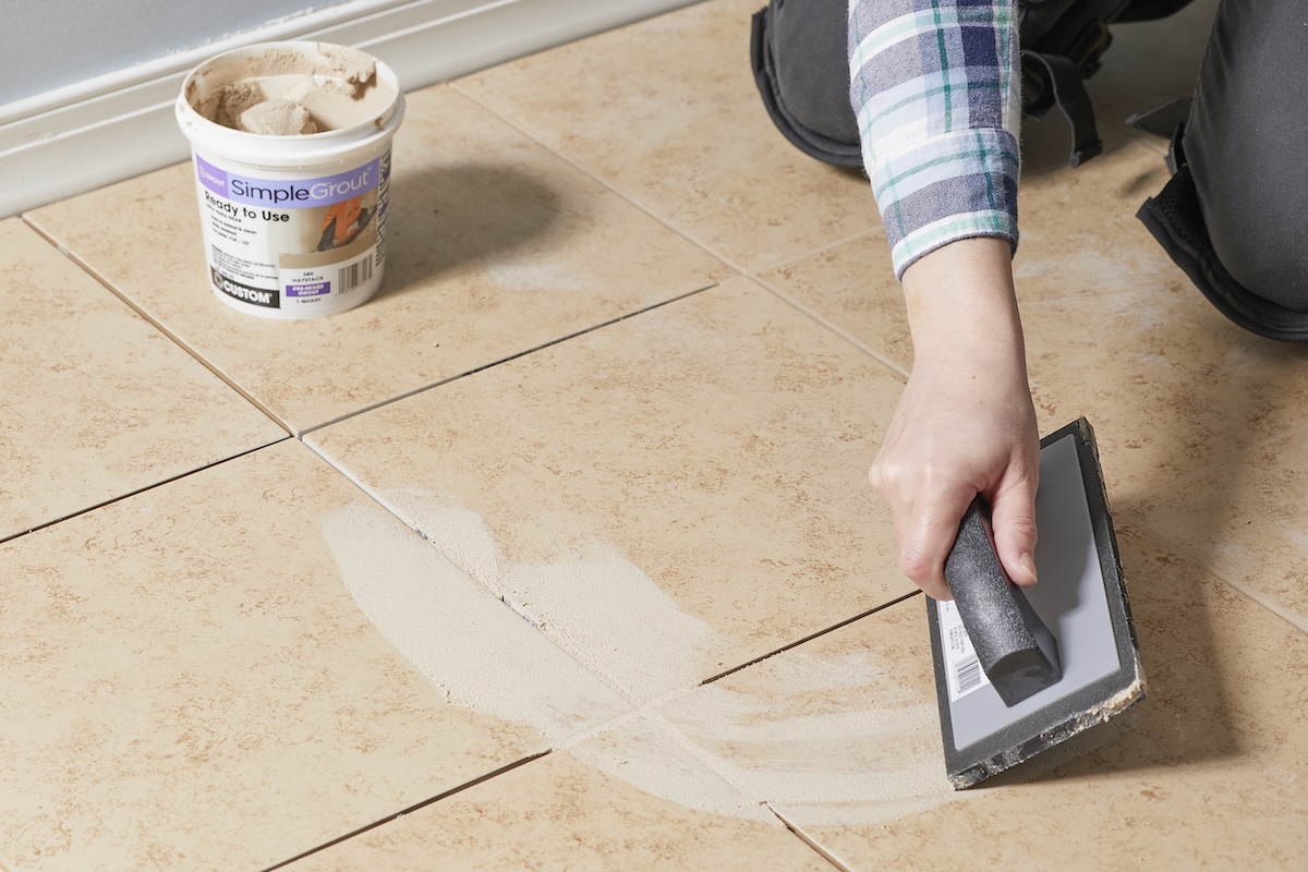 Woman uses grout float to apply grout in an arc across beige floor tiles.