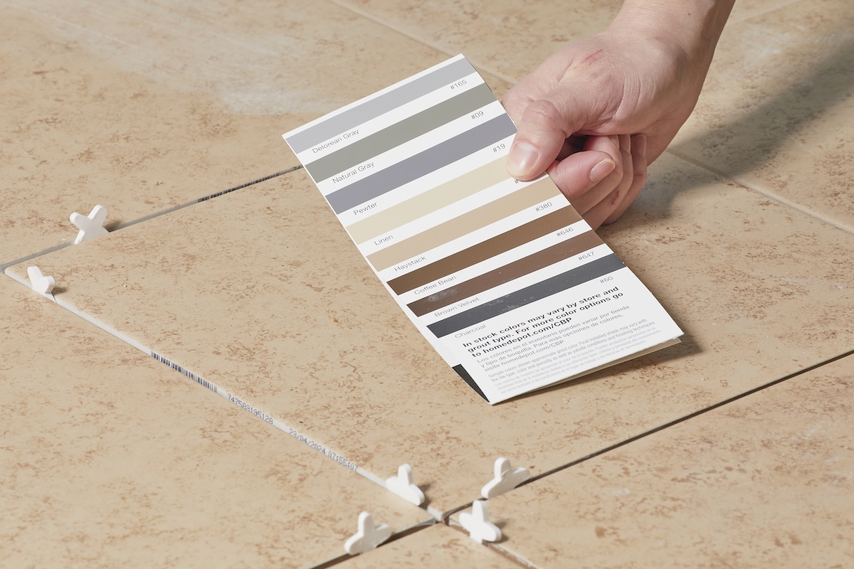 Person holds swatch of grout colors near beige tile floor with spacers between tiles.