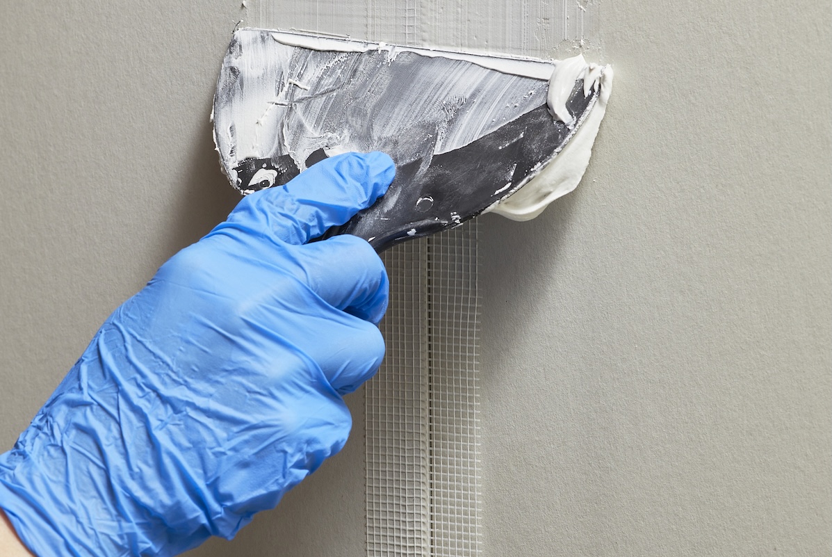Person wearing plastic gloves uses puttty knife to apply joint compound to drywall tape on drywall. 