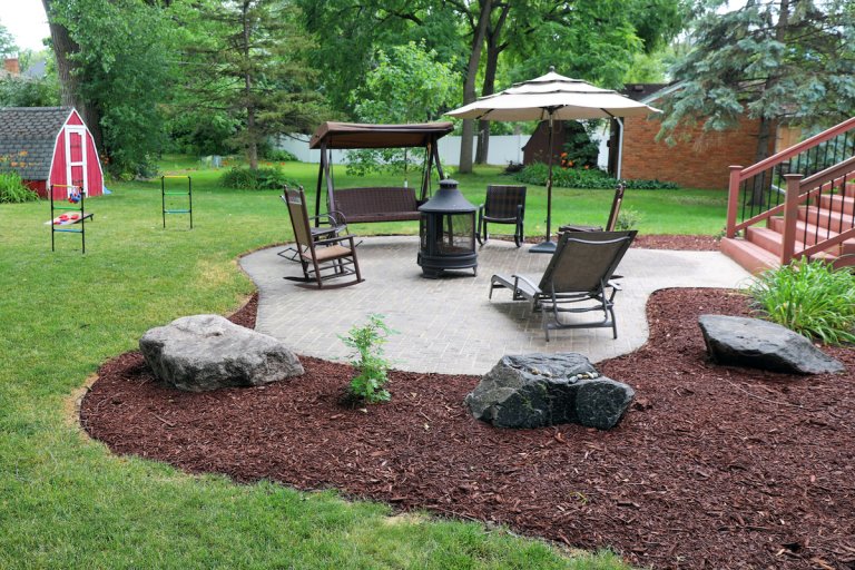 A backyard's concrete patio is surrounded by a mulched garden or plants and large rocks.