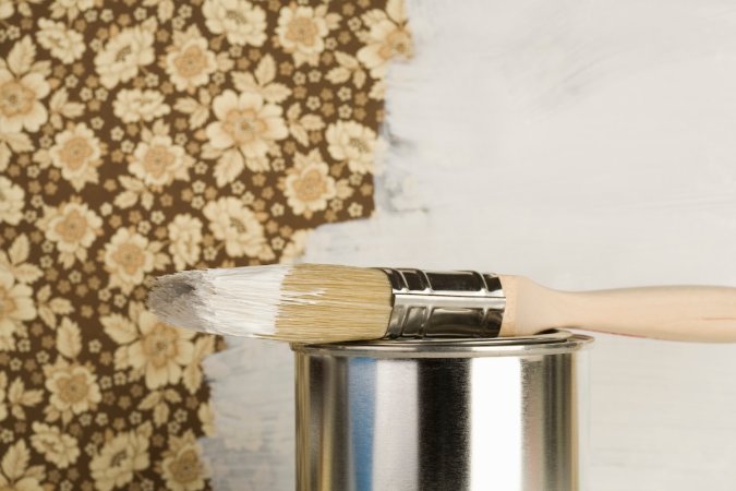 A can of white paint and paintbrush in front of a half-painted wall of brown floral wallpaper.