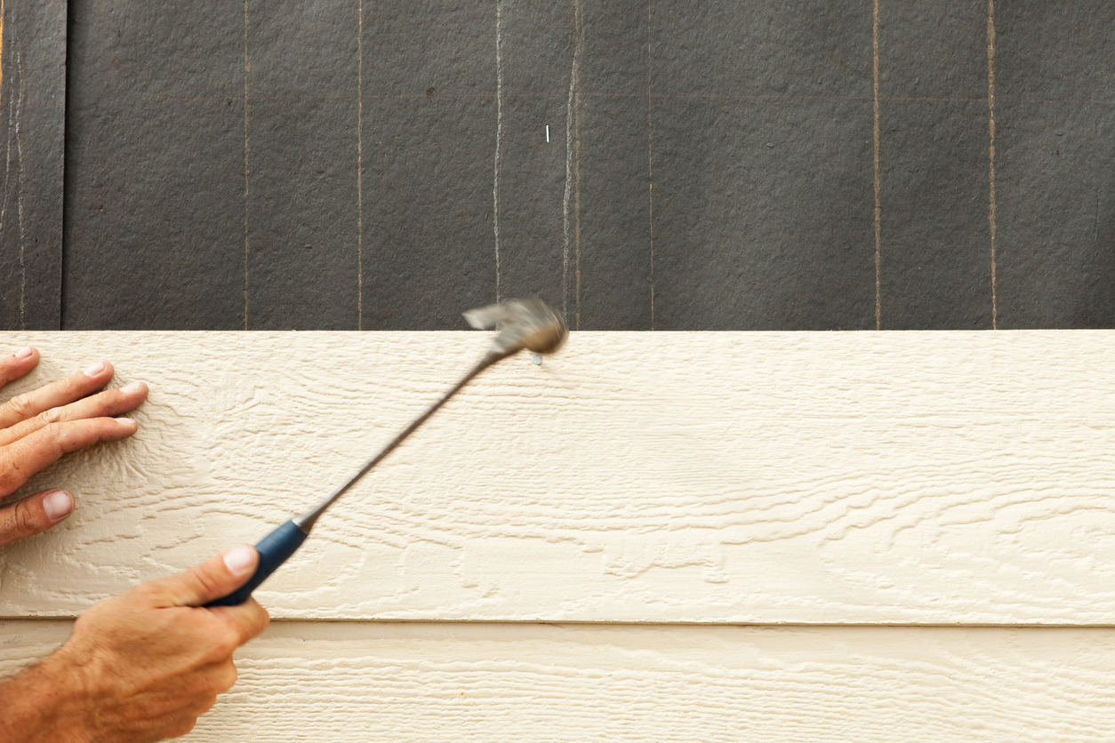 A construction worker, using a hammer, is installing siding onto a tar paper covered exterior house wall. There is some motion blur in the hammer and the nail head is visible next to the hammer head.