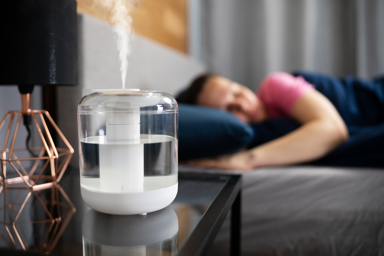 A portable air humidifier sits on a bedside table next to a black lamp while woman in pink shirt sleeps in the background.