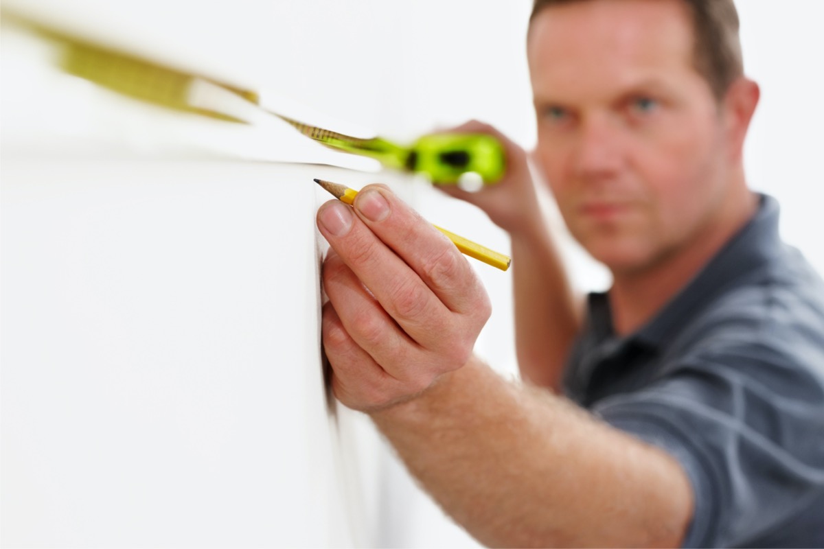 Man uses a tape measure to measure horizontally across a wall, marking a spot with a pencil.