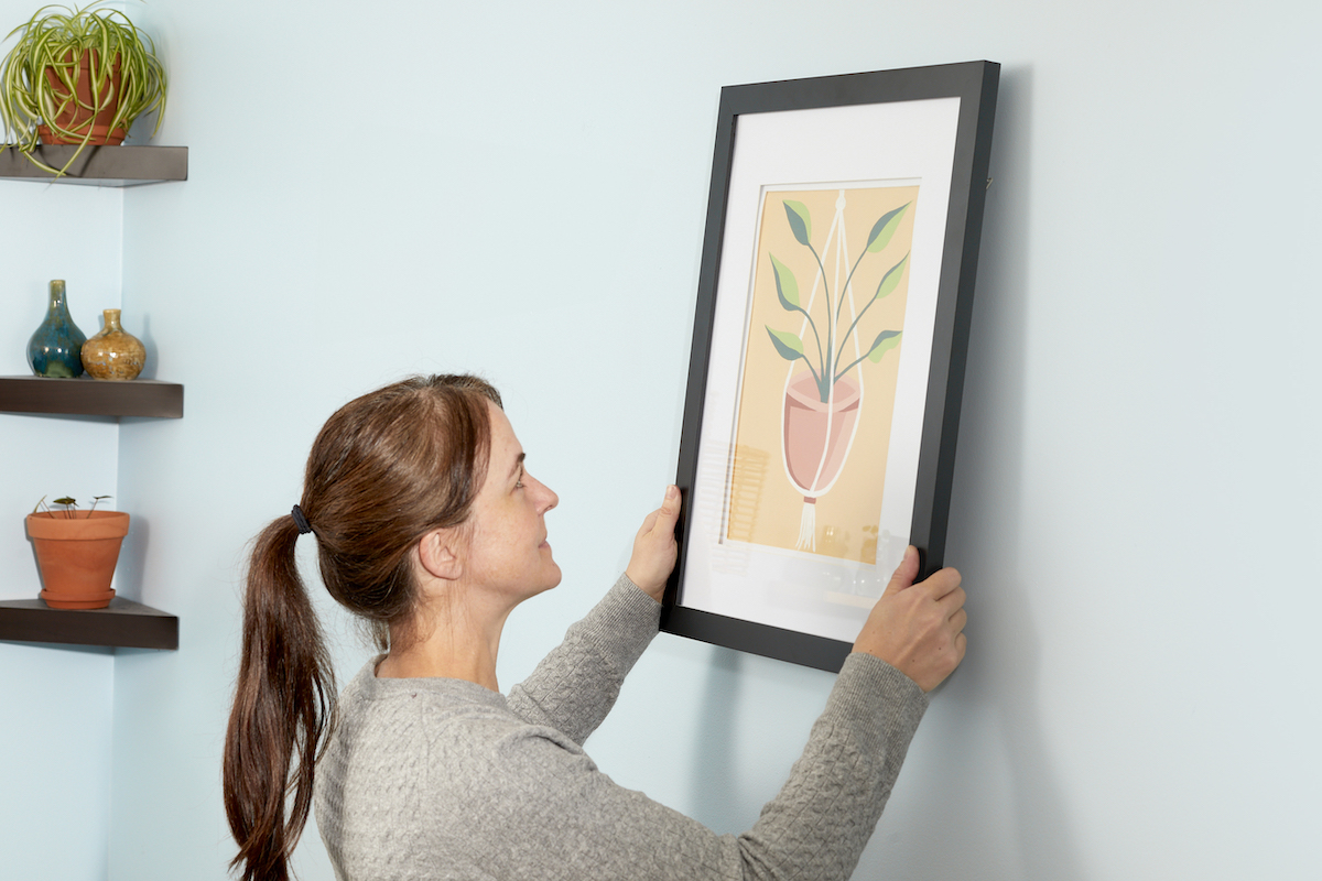 Woman hangs a framed piece of artwork on a light blue wall, with floating shelves in the corner of the room.