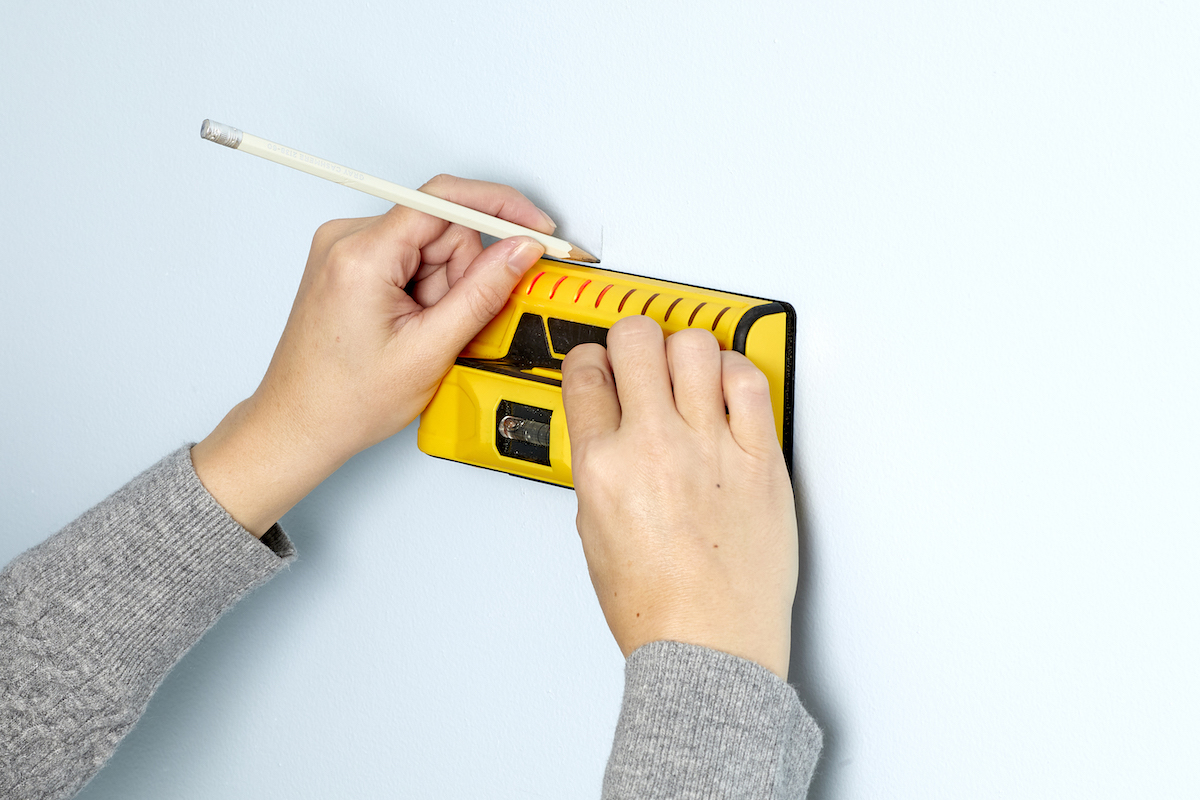 Woman uses a stud finder on a wall, and marks the wall with a pencil.