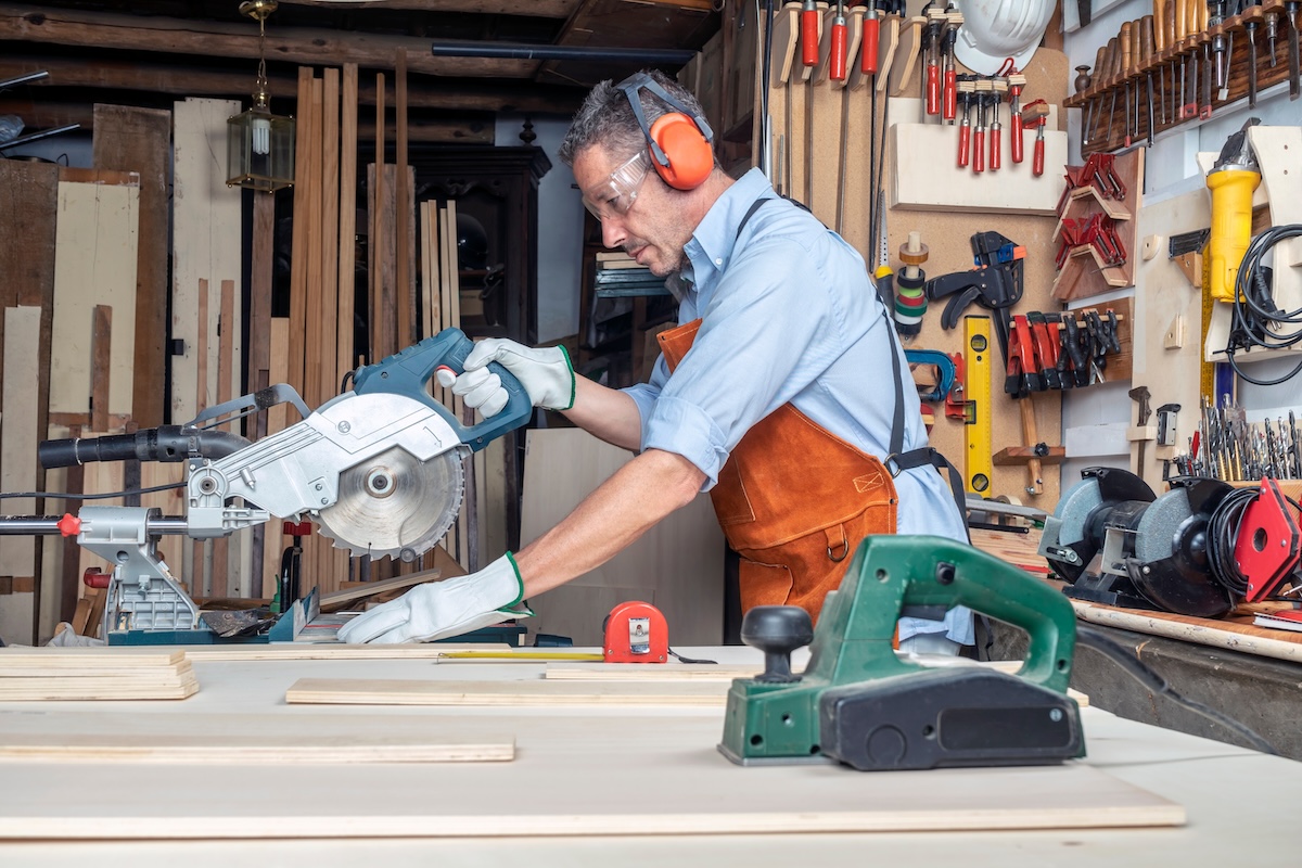 A woodworker with a leather apron and safety gear uses a miter saw in a wood shop.