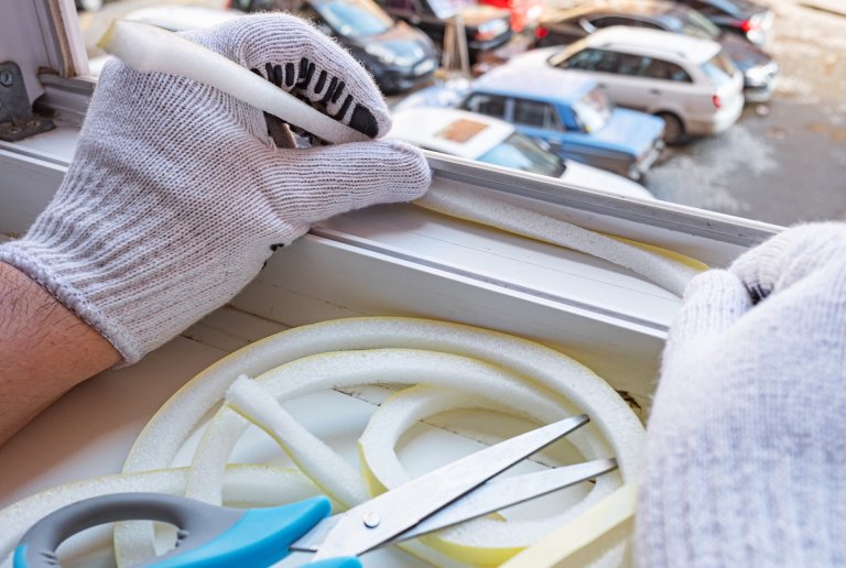 Person wearing work gloves prepares to add foam rubber insulation to window overlooking a parking lot.