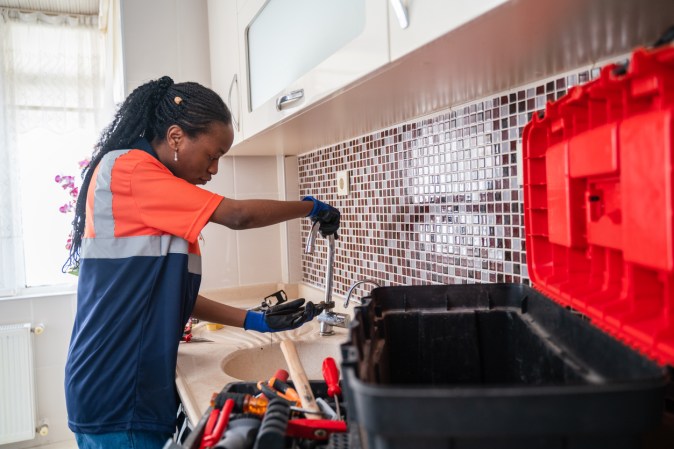 Female plumber with toolbox open troubleshooting water pressure at the kitchen sink.