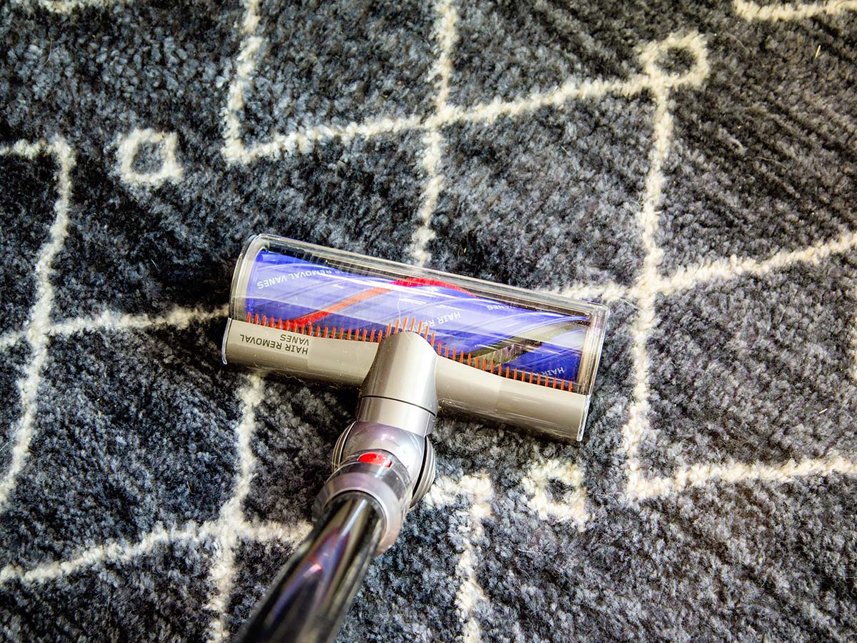A Dyson stick vacuum in use cleaning a grey and white patterned rug.