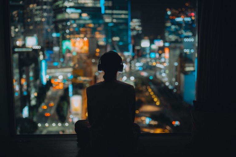 Rear view, silhouette of a woman wearing headphones, looking out window at illuminated city at night.