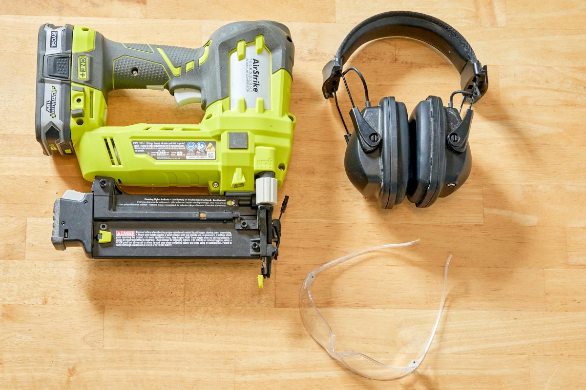 A nail gun, protective headphones, and safety goggles on a wood table.