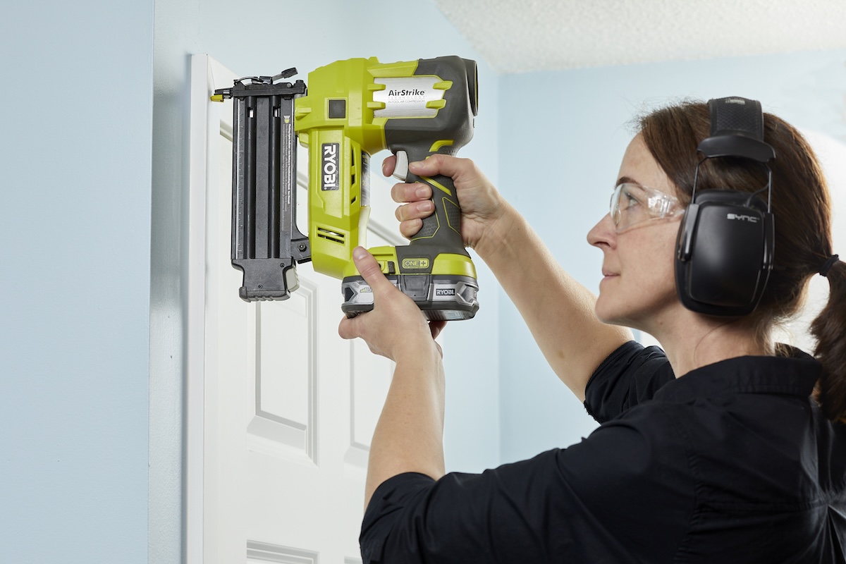 Woman wearing safety goggles positions nail gun on door trim.