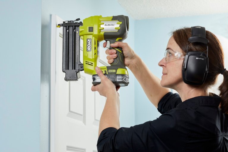 Woman wearing safety goggles positions nail gun on door trim.